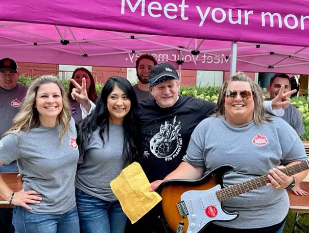 three people standing in a line in front of a tent facing the camera, one holding the guitar that was given away